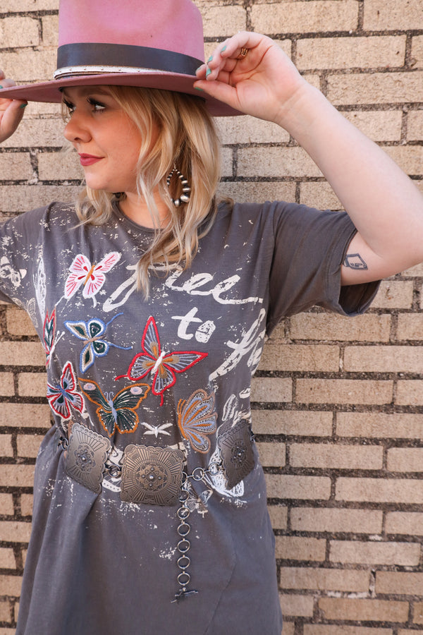 Woman wearing grey t-shirt dress features a bold script that reads "Free to fly" and is adorned with embroidered butterflies and splatter paint. Embrace your unique and fearless spirit with this effortlessly cool and artistic dress.