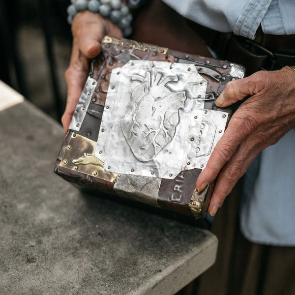 Human hands holding a box with a lid comprised of a mixture of metals and a human heart image displayed on the top