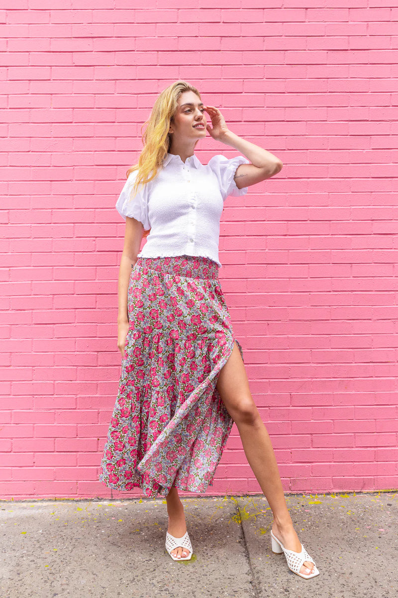 Woman in a white blouse and floral skirt standing against a pink brick wall.