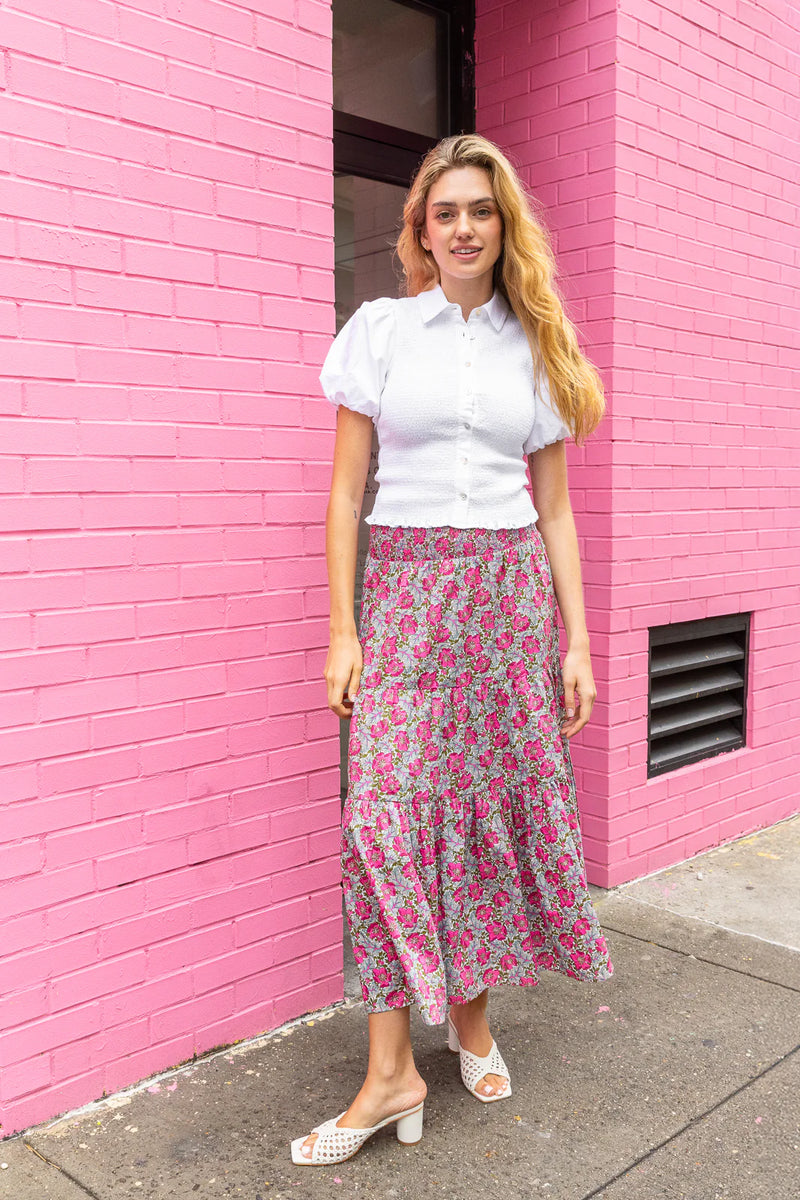 Woman in a white blouse and floral skirt standing in front of a pink brick wall.