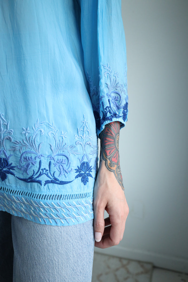 Light blue embroidered top worn by a person with a tattooed arm against a neutral background