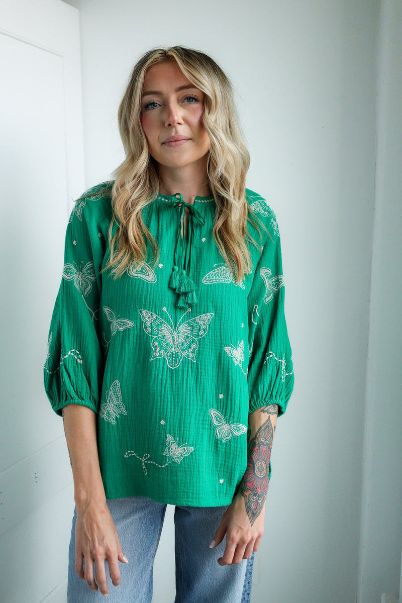 Woman wearing a green blouse with butterfly patterns against a white wall.