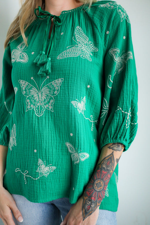 Green blouse with butterfly patterns worn by a person with tattoos on a plain background