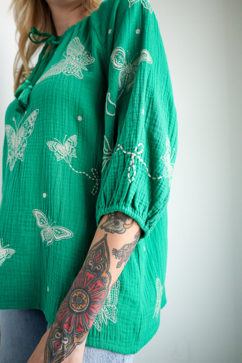 Green patterned top with butterfly design worn by a person with tattoos on a plain background