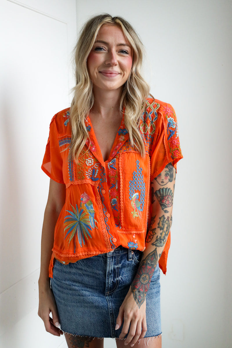 Woman wearing an orange embroidered blouse and denim skirt against a white background
