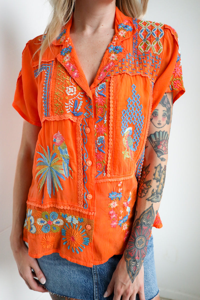 Person wearing an orange embroidered blouse with blue patterns on a plain background