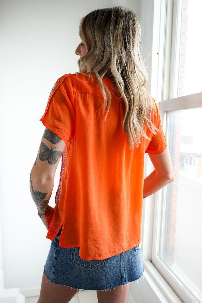 Woman wearing an orange top and denim skirt standing by a window.