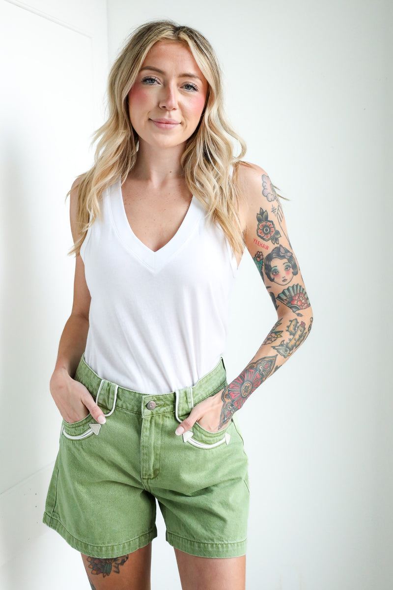 Woman wearing a white tank top and green shorts with tattoos on her arms and legs, standing against a plain background.