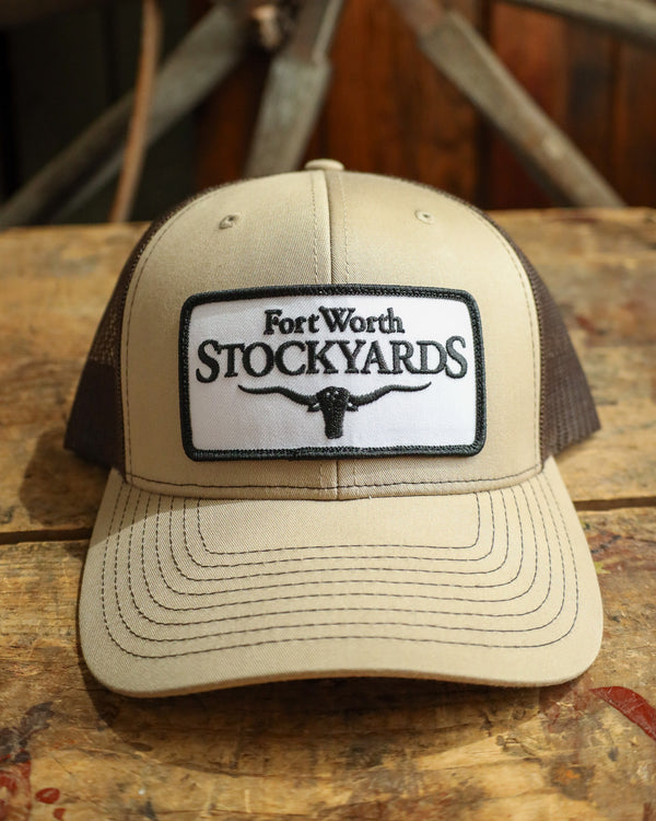 Beige and brown trucker hat with Fort Worth Stockyards logo on a wooden surface