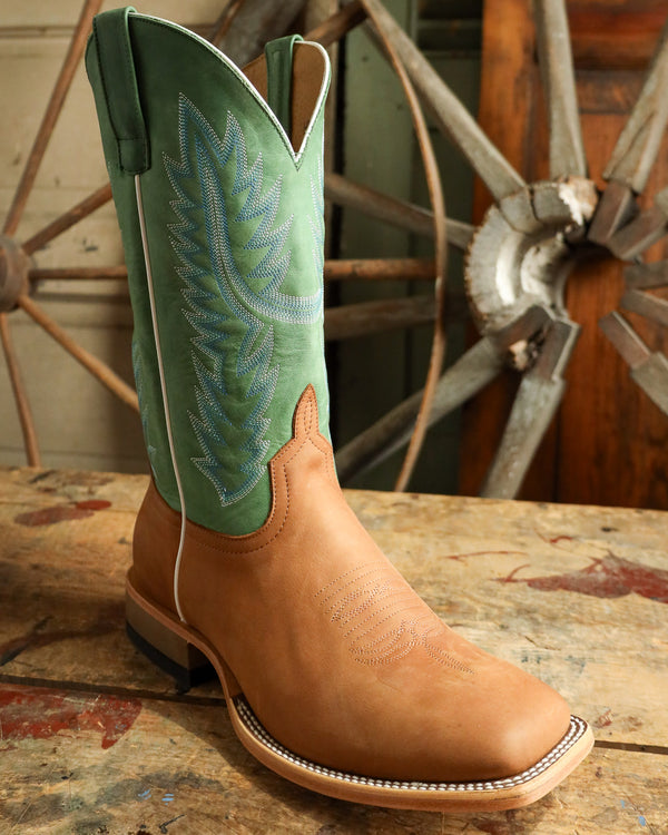 Brown cowboy boot with green embroidered details on a wooden surface with a rustic background.