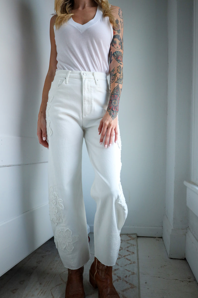 Person wearing a white sleeveless top and white pants with lace details, standing against a plain wall.