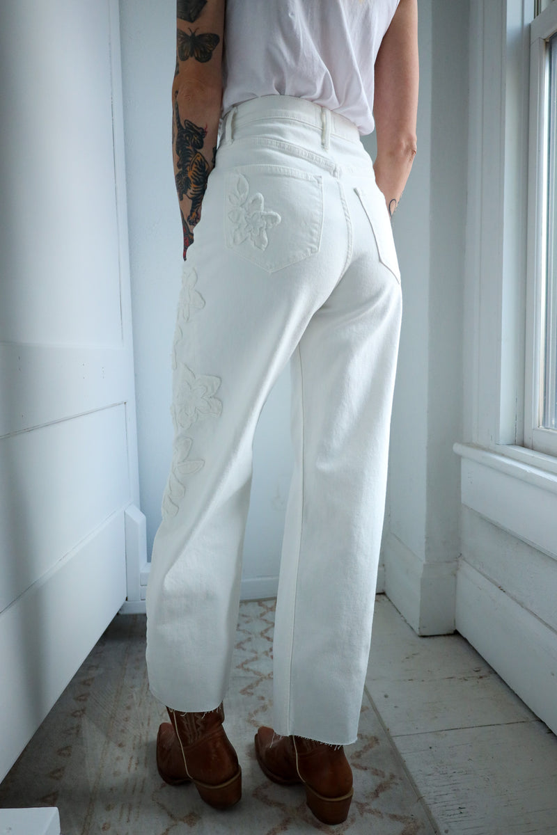 Person wearing white pants with floral embroidery in a room with white walls and a tiled floor.