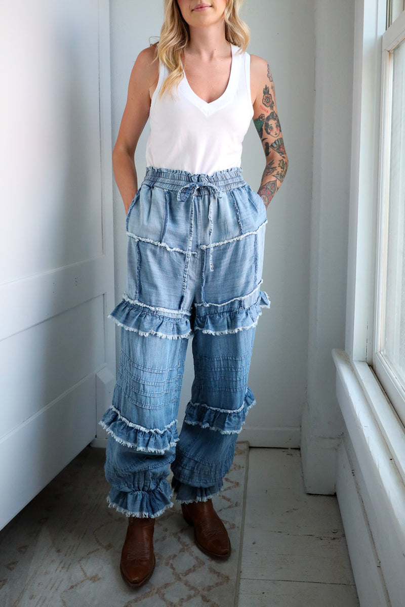 Woman wearing a white tank top and blue tiered denim pants in a room with a window.