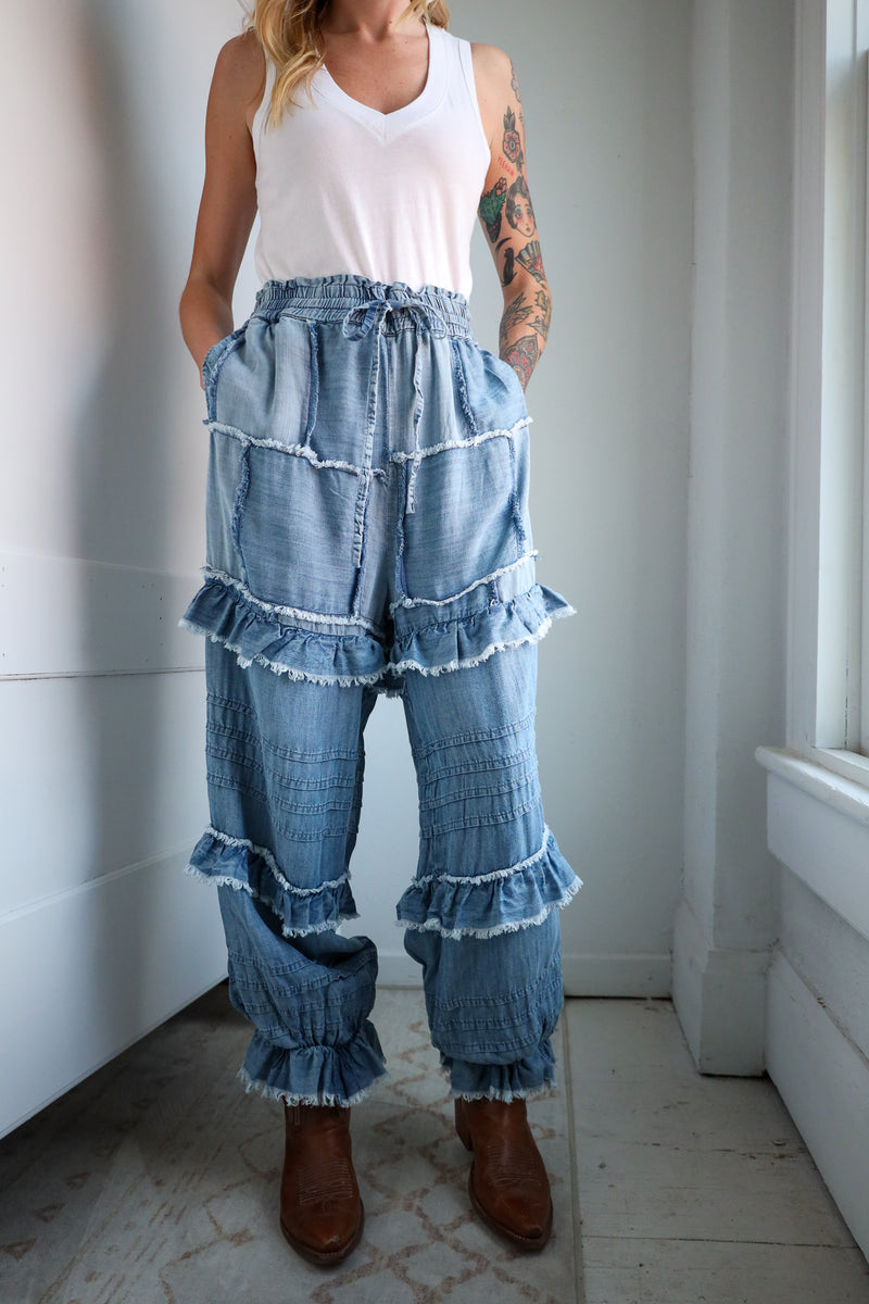 Person wearing a white tank top and blue denim pants with ruffles in a minimal indoor setting.