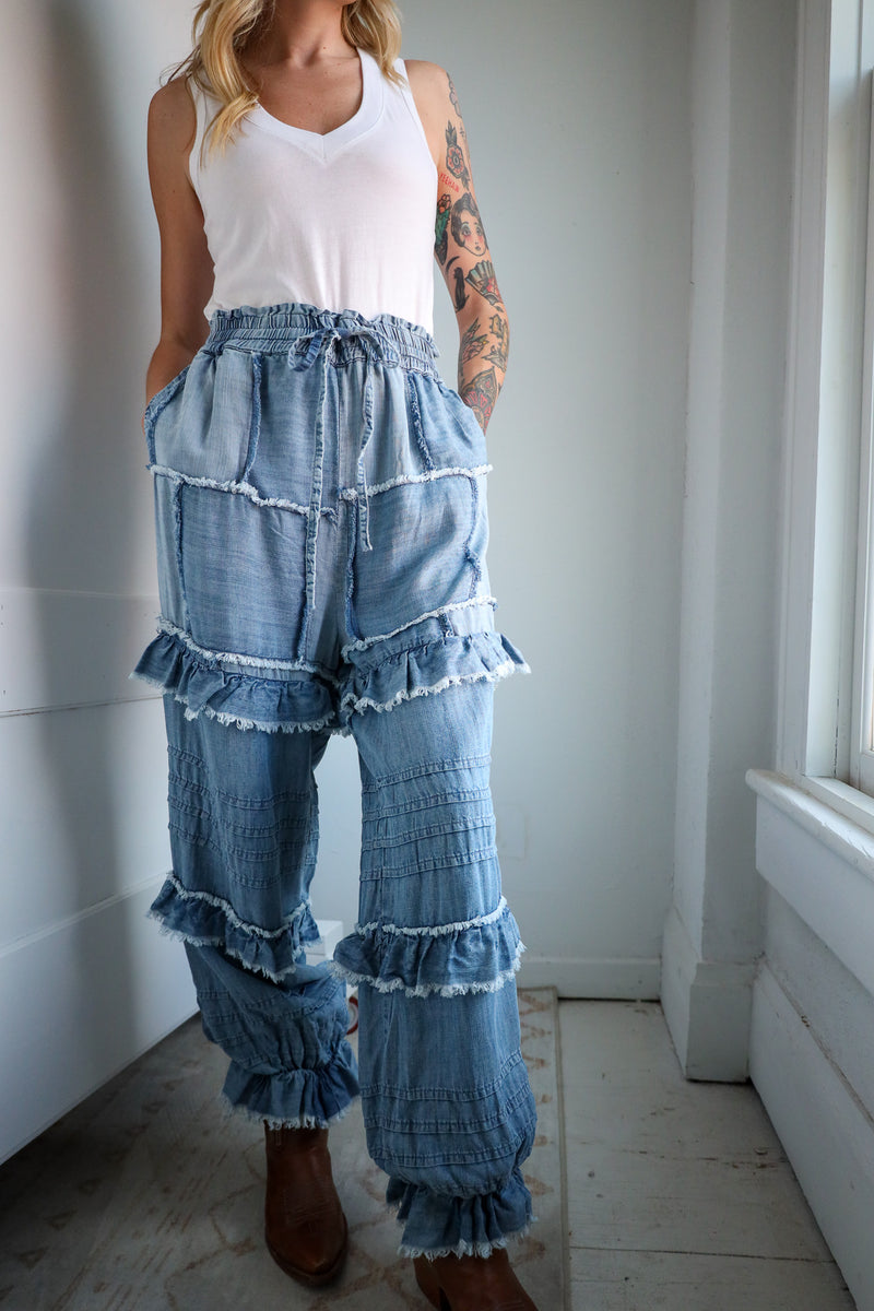 Woman wearing a white tank top and blue tiered denim pants in a room with white walls and tiled floor.