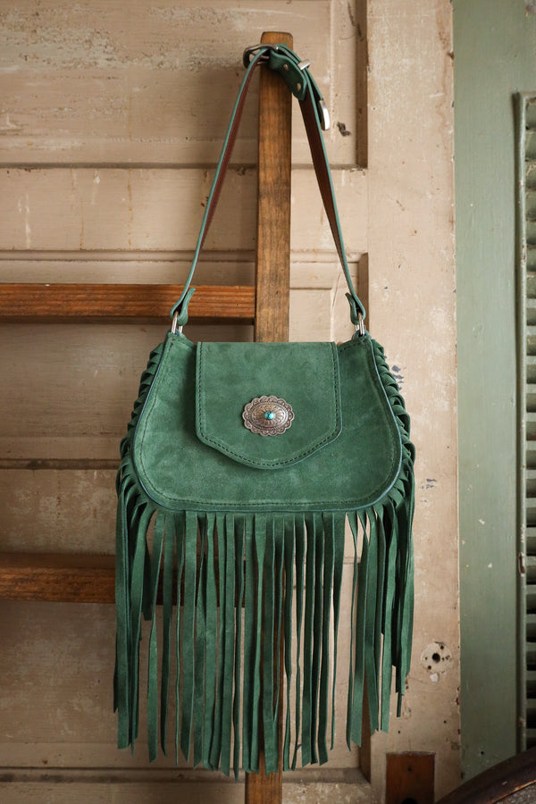 Green suede handbag with fringe and a decorative clasp on a wooden stand.
