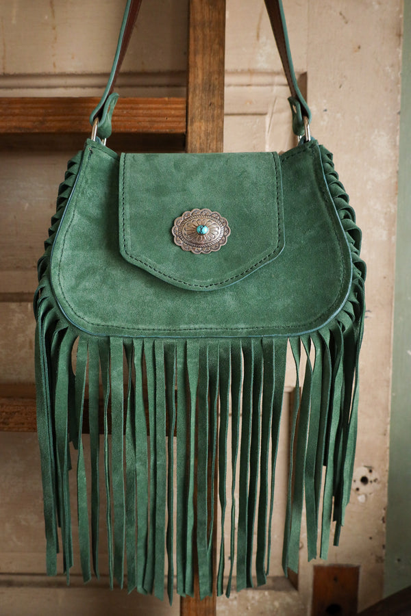 Green suede handbag with fringe and a central clasp on a wooden stand.
