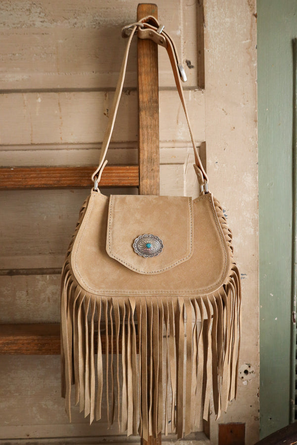 Beige suede handbag with fringe and a concho on a wooden surface.