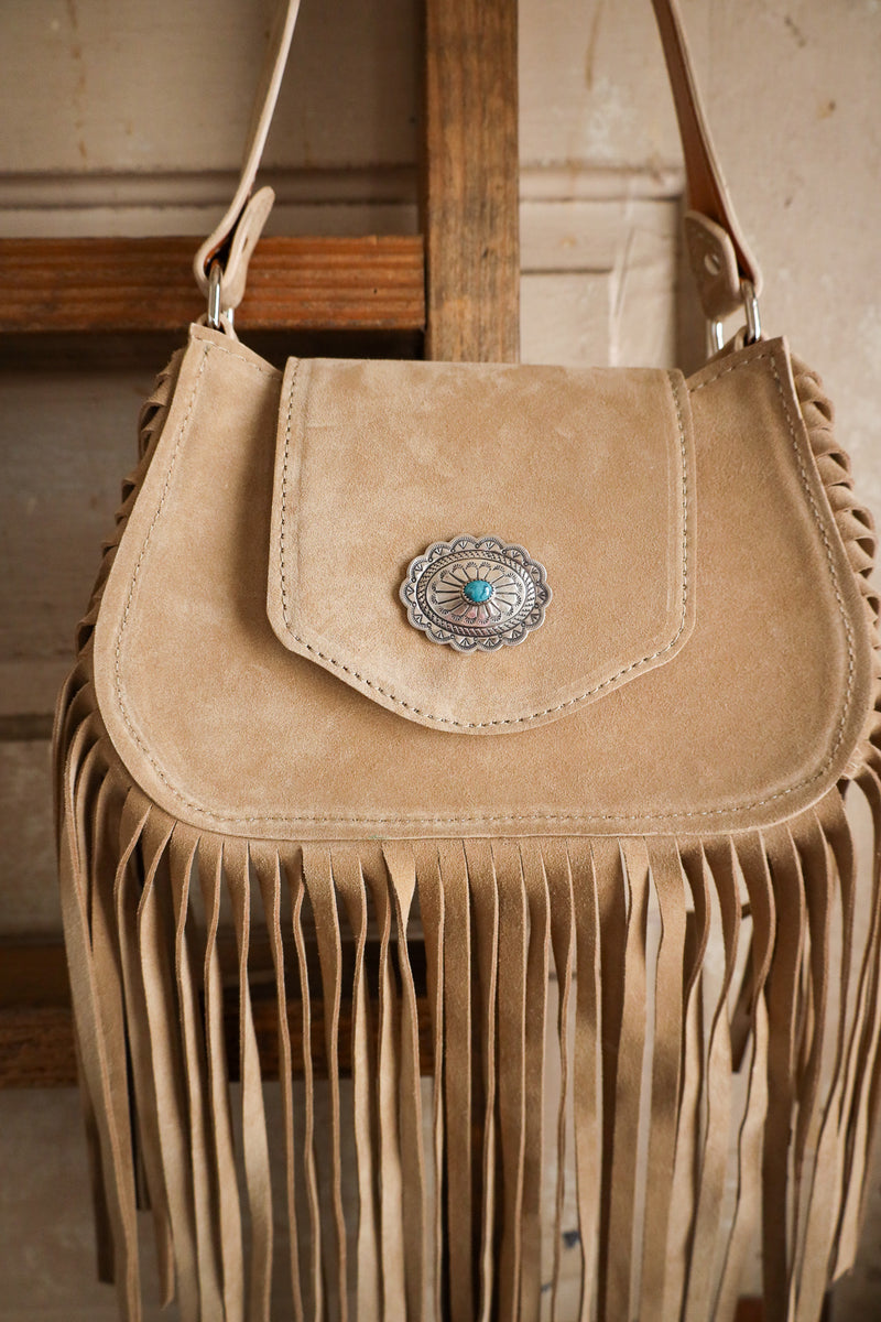Beige suede handbag with fringe and a decorative clasp on a wooden surface.