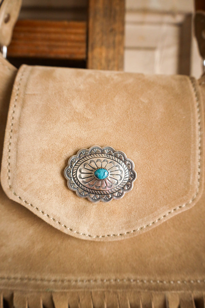 Close-up of a tan leather bag with a silver and turquoise concho on a wooden surface.
