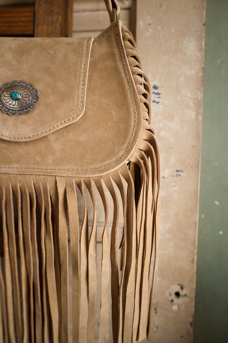 Brown suede handbag with fringe details and a decorative clasp on a textured wall.