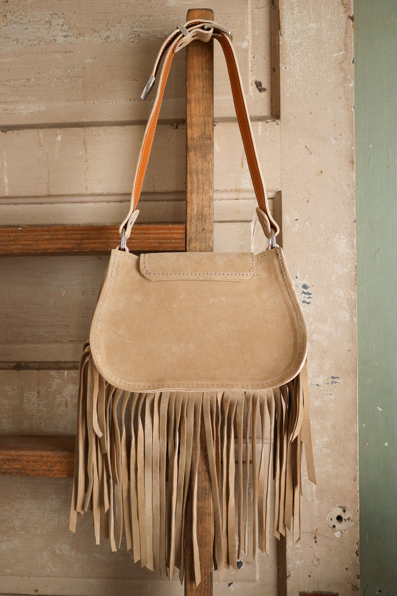 Beige suede handbag with fringe hanging on a wooden surface.