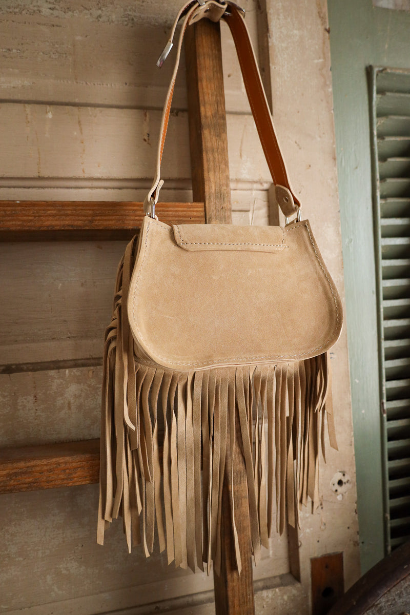 Beige suede handbag with fringe hanging on a wooden post against a rustic background.