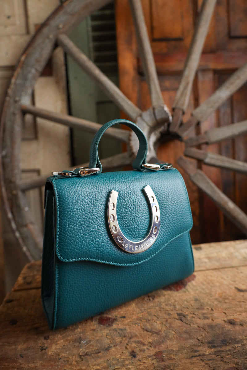 Teal handbag with a horseshoe clasp on a wooden surface with a rustic wheel background