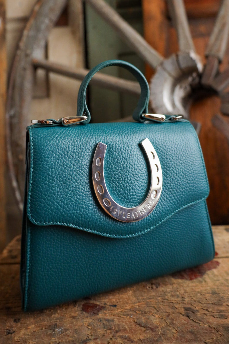 Teal handbag with a silver buckle on a wooden surface with a rustic wheel in the background