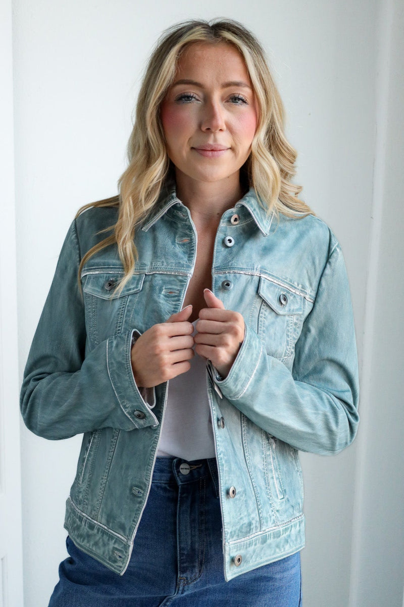 Woman wearing a denim jacket and jeans indoors against a white wall.