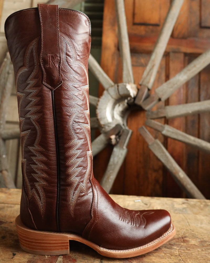 Brown leather cowboy boot with intricate patterns on a wooden surface with a rustic background.