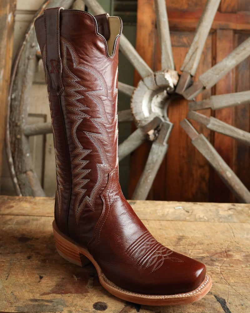Brown cowboy boot with intricate designs on a wooden surface with a rustic wheel background