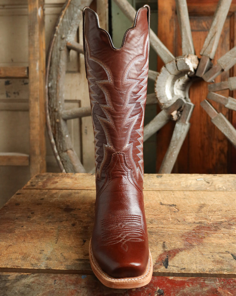 Brown cowboy boot with intricate design on a wooden surface with a wagon wheel in the background