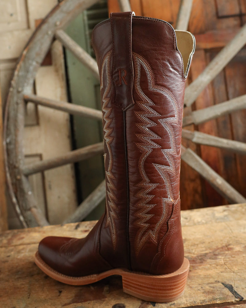 Brown cowboy boot with intricate stitching on a wooden surface with a rustic background