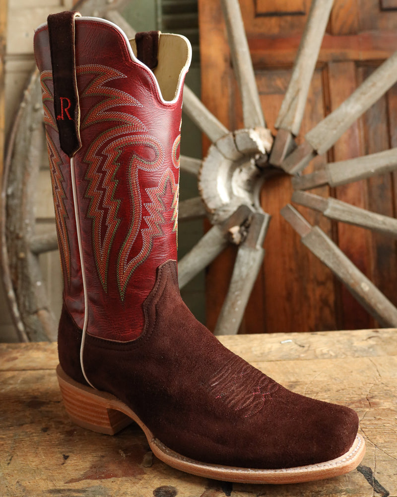 Brown cowboy boot with red accents on a wooden surface with a wagon wheel in the background