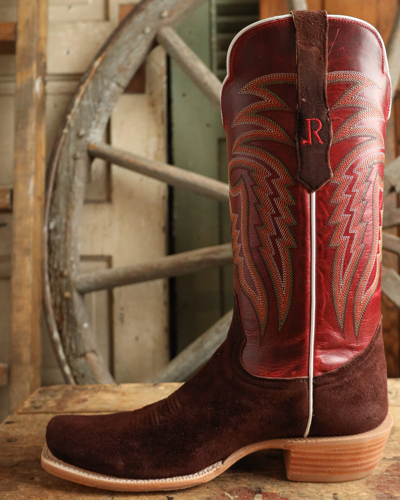 Brown and red cowboy boot with intricate design and 'R' logo, placed on a wooden surface with a rustic background.