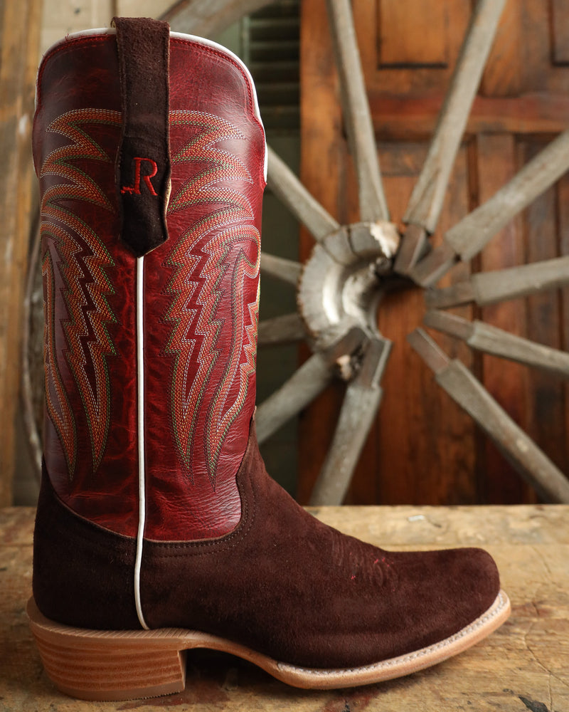 Brown and red cowboy boot with intricate design in front of a wooden wagon wheel.