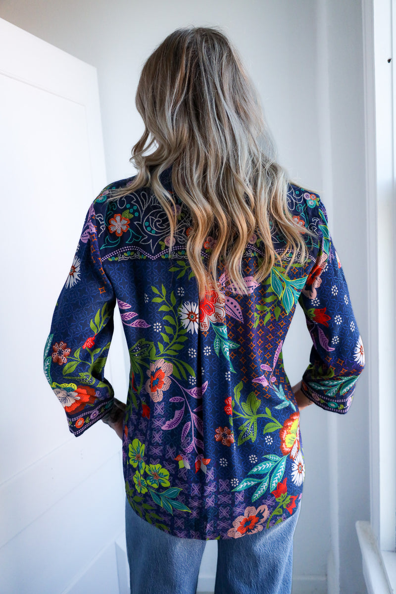Person wearing a colorful floral shirt with long hair, standing against a white wall.