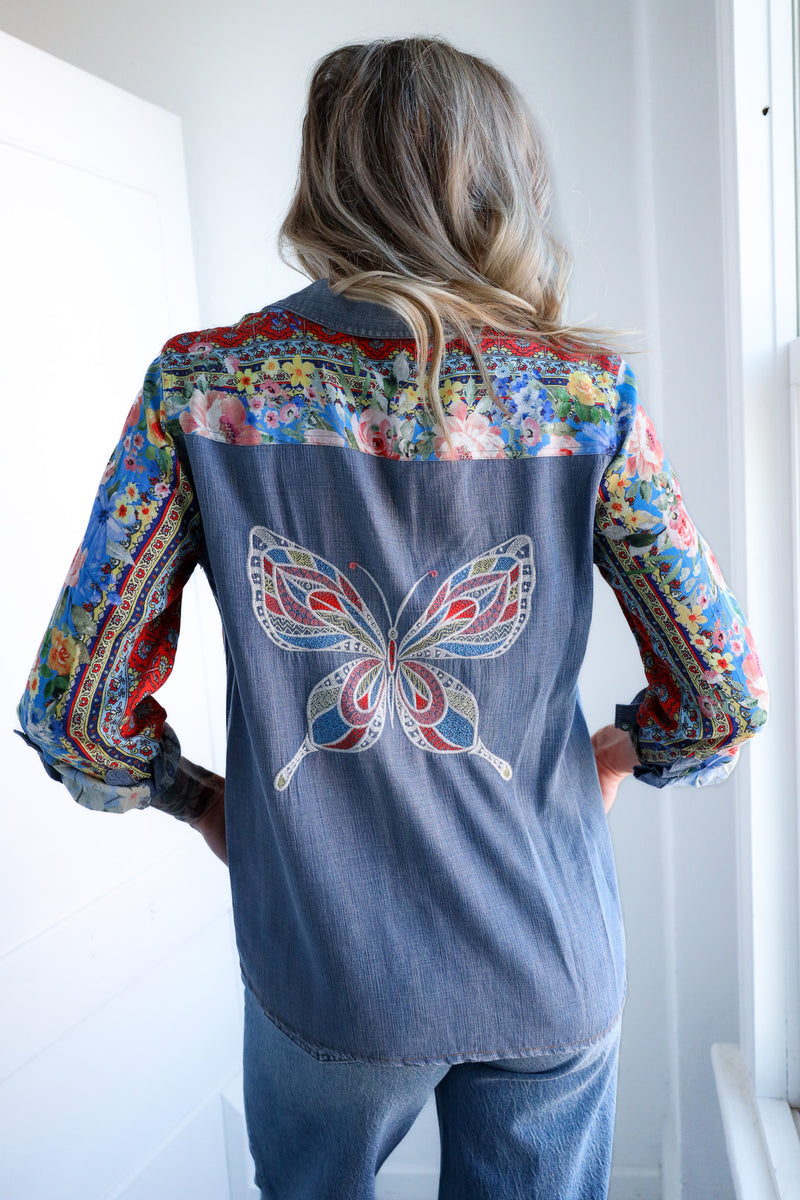 Woman wearing a denim shirt with a colorful butterfly design on the back, standing against a white wall.