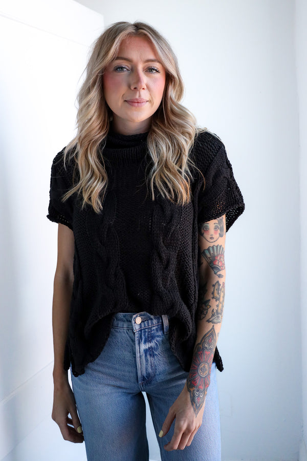 Woman wearing a black cable knit sweater and blue jeans against a white background