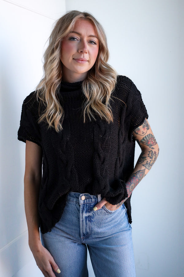 Woman wearing a black cable knit sweater and blue jeans against a white background