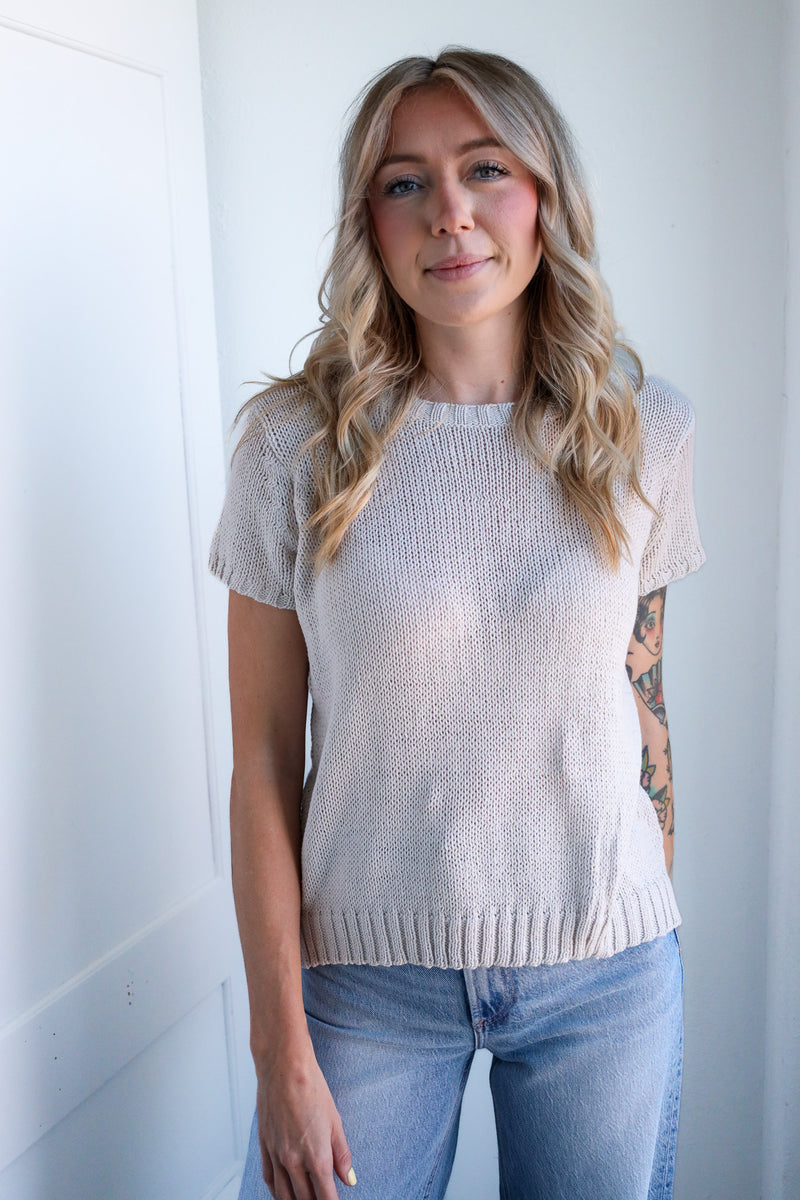 Woman wearing a light beige sweater and blue jeans against a white wall