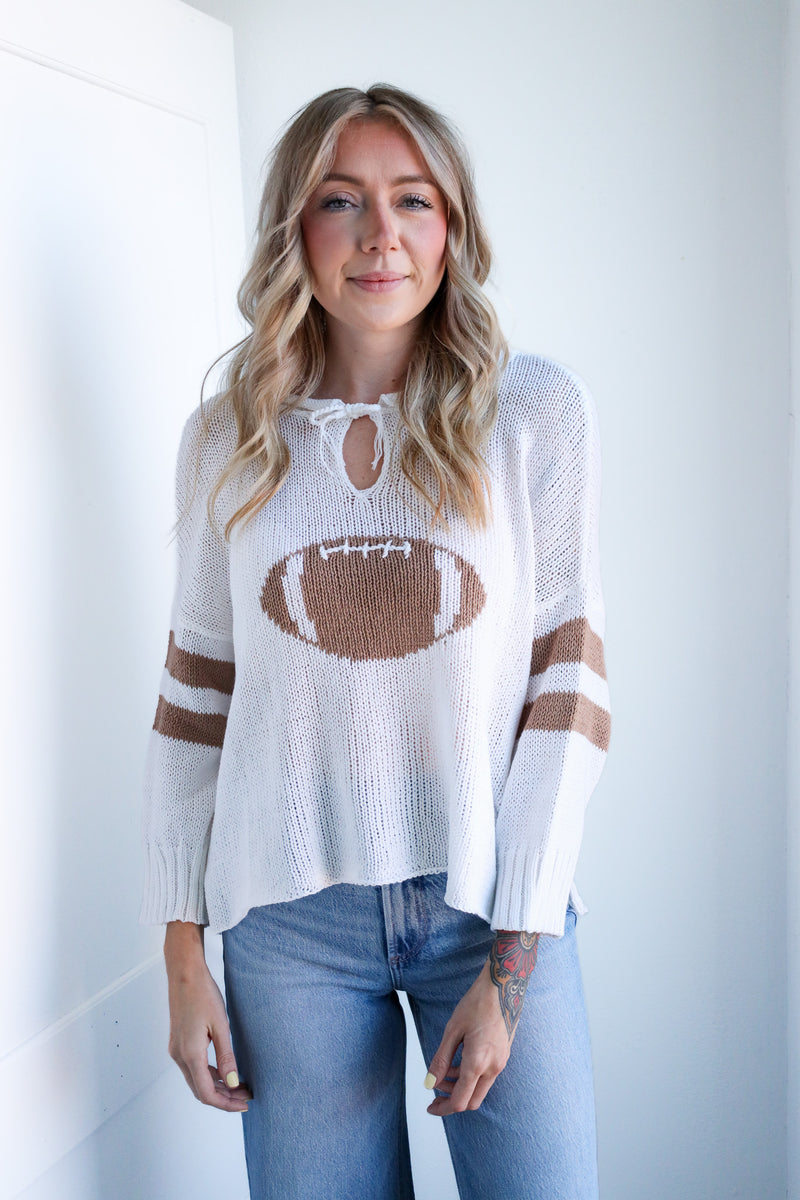 Woman wearing a white sweater with a football design and blue jeans indoors.