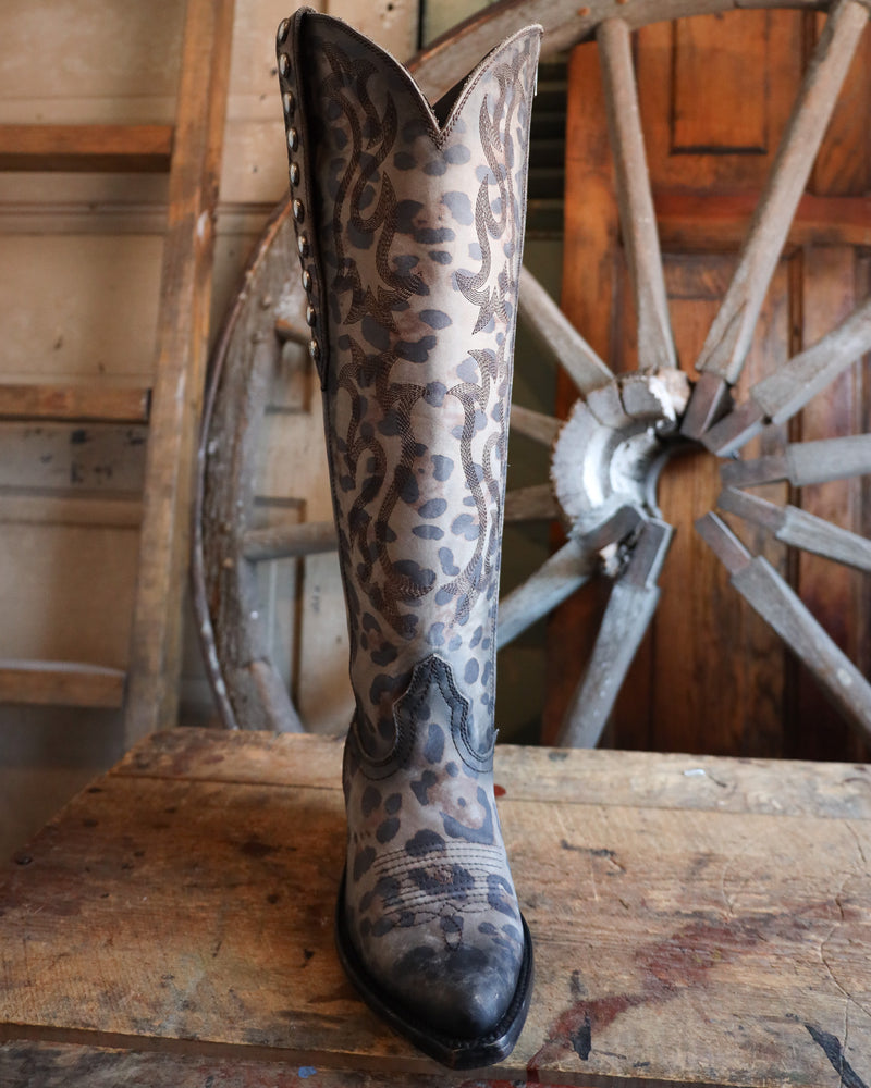 Cowboy boot with leopard print pattern on a wooden surface with a wagon wheel in the background