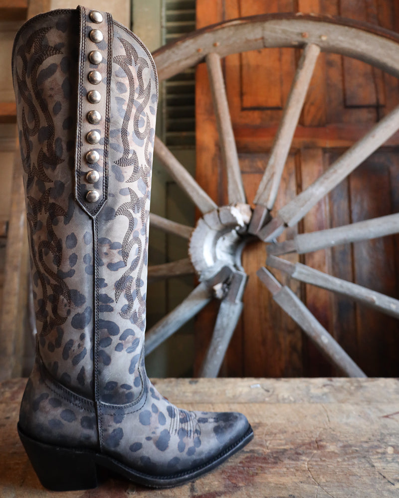 Leopard print cowboy boot with studded details in front of a wooden wagon wheel.