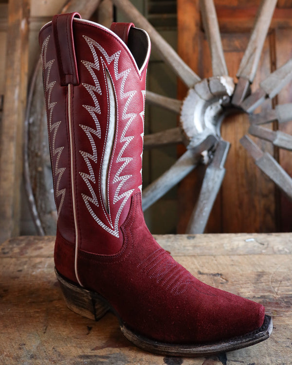 Red cowboy boot with white stitching on a wooden surface with a wheel in the background