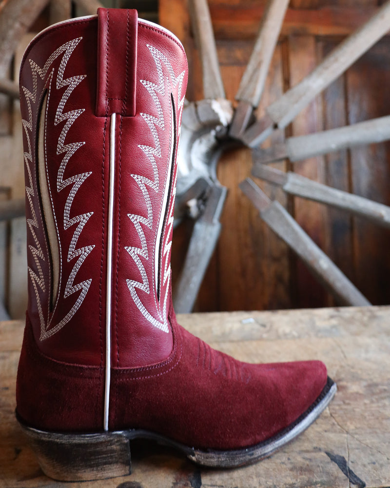 Red cowboy boot with white stitching on a wooden surface with a wagon wheel in the background