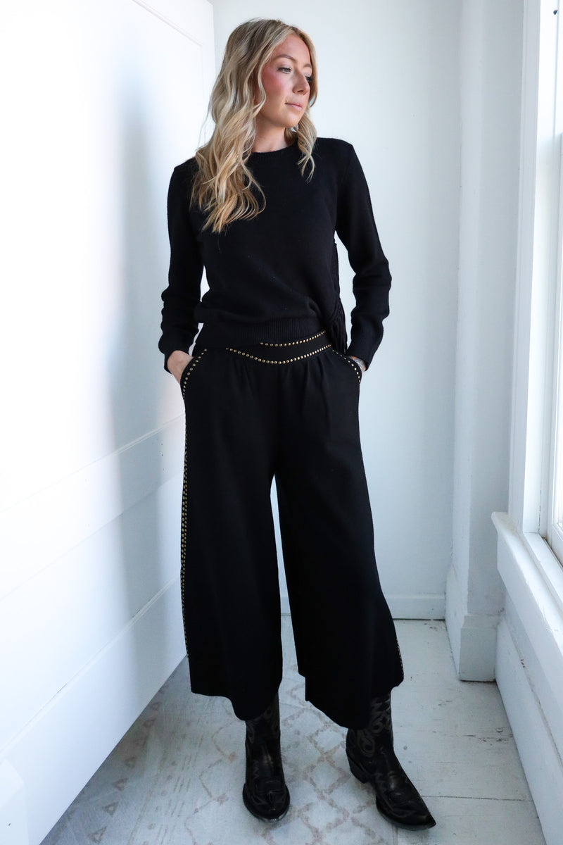 Woman wearing a black outfit with wide-leg pants in a minimalistic room.
