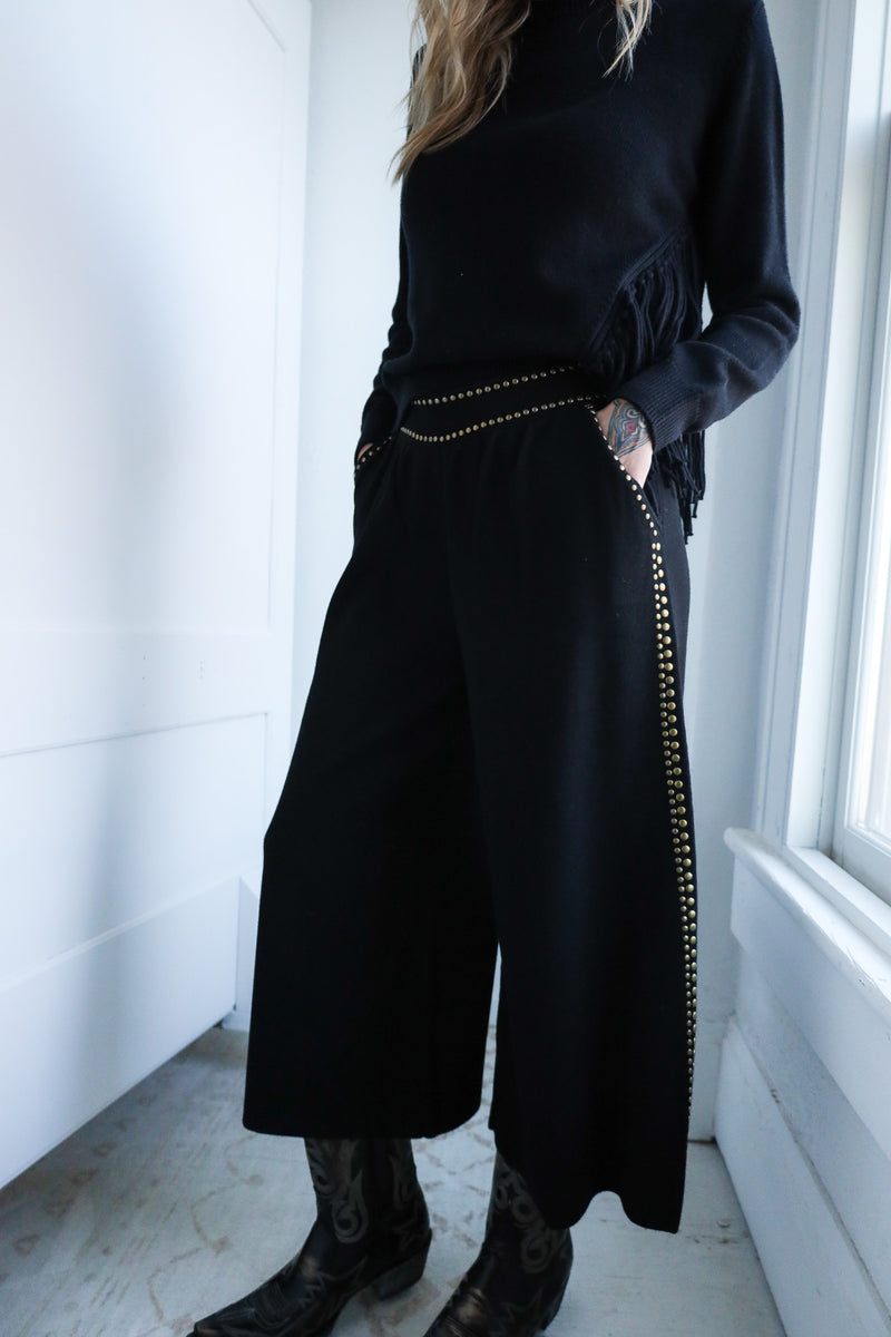 Person wearing a black outfit with wide-leg pants and a high-neck top, standing in a room with a window.