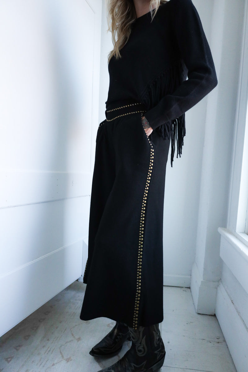 Person wearing a long black coat with fringe details in a minimalistic room.